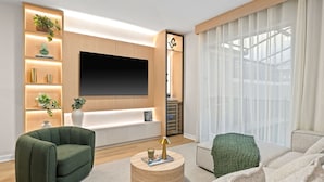 A modern living room with a flat-screen TV, built-in shelves, a sofa, and a large window with sheer curtains.