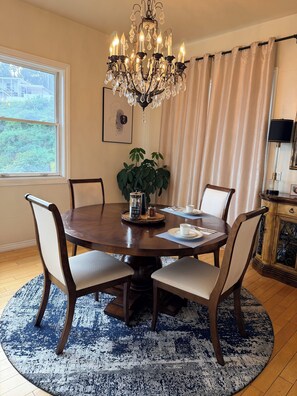 A dining room with a round table, four chairs, a chandelier, a potted plant, and a window with curtains.