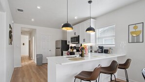 A modern kitchen with white cabinetry, a central island with bar stools, pendant lighting, and a view of the adjacent living area. - Minutes to Downtown Spacious 4BR 3BA Historic Gem (San Antonio)