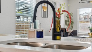 A modern kitchen with a stainless steel sink, a black faucet, and a soap dispenser. There is a window with blinds, a vase with flowers, and a plant on the counter. - Minutes to Downtown Spacious 4BR 3BA Historic Gem (San Antonio)