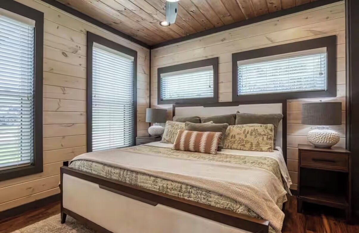 A bedroom with a wooden ceiling, a bed with a beige and brown patterned bedspread, two bedside lamps, and a nightstand with a decorative bowl.