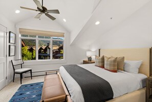 A bedroom with a bed, a chair, a bench, a window with a view, a ceiling fan, and a nightstand with a lamp. - Modern Big Sky Retreat  | Hot tub + Shared Pool (Big Sky)