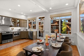 A modern kitchen with stainless steel appliances, a dining area with a long table set for six, and large windows offering a view of a wooden house and greenery. - Modern Big Sky Retreat  | Hot tub + Shared Pool (Big Sky)
