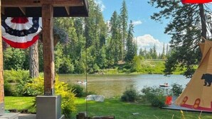 A wooden structure with a flag, a river, and a tent with a bear design. - New! Luxury Swan River Waterfront Home | Reclaimed Timber, Fire Pit & Mountain Views (Bigfork)