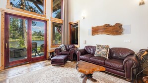 A spacious living room with a large wooden-framed window, a leather sofa, a wooden coffee table, and a rustic wall art piece. - New! Luxury Swan River Waterfront Home | Reclaimed Timber, Fire Pit & Mountain Views (Bigfork)