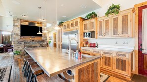 A modern kitchen with a large island, wooden cabinets, and a stone accent wall. - New! Luxury Swan River Waterfront Home | Reclaimed Timber, Fire Pit & Mountain Views (Bigfork)