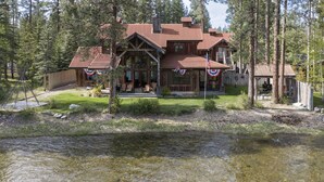 A lodge with a large porch, a covered outdoor seating area, and a river in front. - New! Luxury Swan River Waterfront Home | Reclaimed Timber, Fire Pit & Mountain Views (Bigfork)