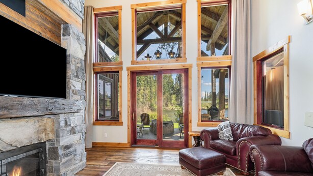 A stone fireplace, leather sofas, and large windows with a view of trees. - New! Luxury Swan River Waterfront Home | Reclaimed Timber, Fire Pit & Mountain Views (Bigfork)