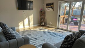 A living room with grey sofas, a patterned rug, a flat-screen TV, a wooden side table, and a sliding glass door leading to an outdoor patio. - Charming 4-bedroom 2.5 Bath one level house with garage 7 miles from NRG stadium (Houston)