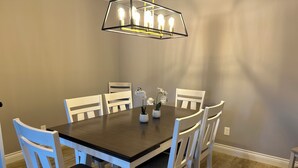 A dining area with a round table, six white chairs, a modern chandelier, and a vase with flowers. - Charming 4-bedroom 2.5 Bath one level house with garage 7 miles from NRG stadium (Houston)