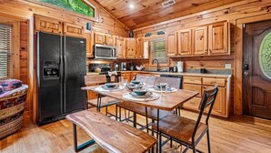 Interior de una cabaña de madera con una mesa de comedor preparada para cuatro personas, un refrigerador negro, un microondas y un lavamanos.