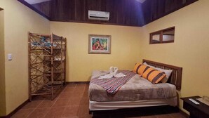 Un dormitorio con una cama, cabecera de madera, aire acondicionado de pared, un cuadro enmarcado y un tendedero.