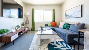 A modern living room with a grey sofa, a coffee table, a TV mounted on the wall, and a bookshelf with plants. - Cozy 2BR W/ Desk • Fast Wi-Fi • Easy Parking (Tucson)