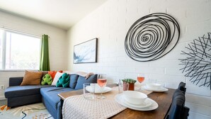 A modern living room with a sofa, a dining table set with plates and glasses, a wall art piece, and a window with curtains. - Cozy 2BR W/ Desk • Fast Wi-Fi • Easy Parking (Tucson)