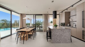 Et moderne køkken med en stor ø, spiseområde med udsigt til en pool, og en rummelig opholdsstue med store vinduer.