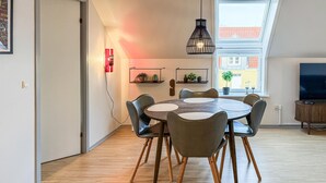 A modern dining area with a round table, six chairs, a pendant light, and a view of a building through a window. - 2 bedroom amazing apartment in Gudhjem (Gudhjem)