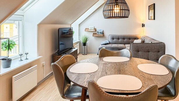 A modern living room with a round wooden dining table, leather chairs, a grey sofa, a television, and a hanging light fixture. - 2 bedroom amazing apartment in Gudhjem (Gudhjem)