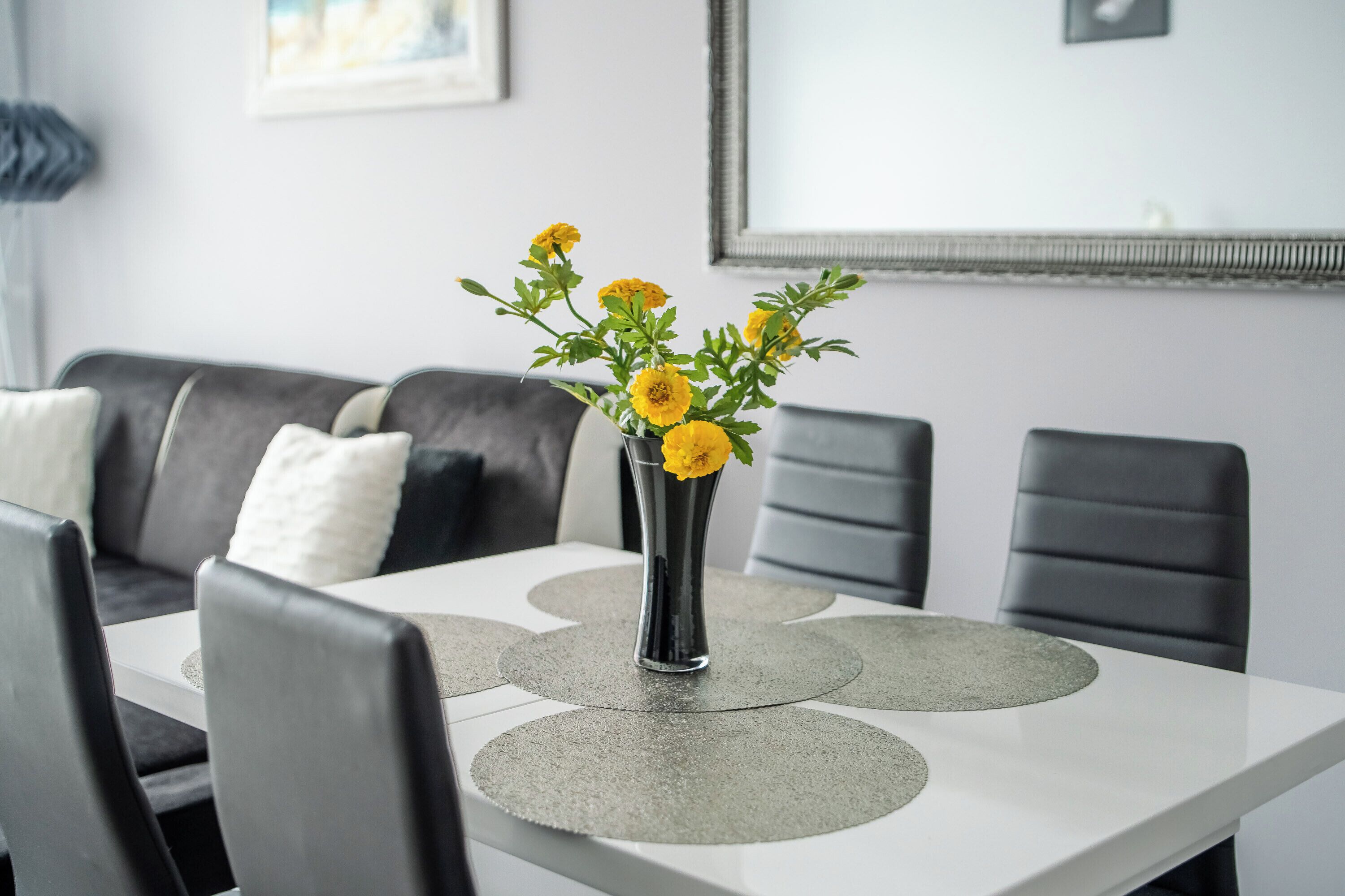 A dining table with a vase of yellow flowers, grey placemats, and black chairs.