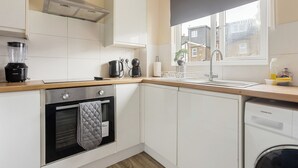 A modern kitchen with white cabinetry, a built-in oven, a dishwasher, and a window with a view of a residential area. - Modern 2BR Flat, 20 mins train to Central London, 7mins to Selhurst Park Stadium (London)