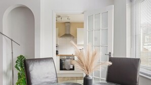 A modern dining area with a glass table, two black chairs, a vase with pampas grass, and a view into the kitchen. - Modern 2BR Flat, 20 mins train to Central London, 7mins to Selhurst Park Stadium (London)