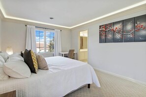 Ein Schlafzimmer mit Bett, Kissen, einem Fenster mit Vorhängen, einem Schreibtisch und einem Bild an der Wand.