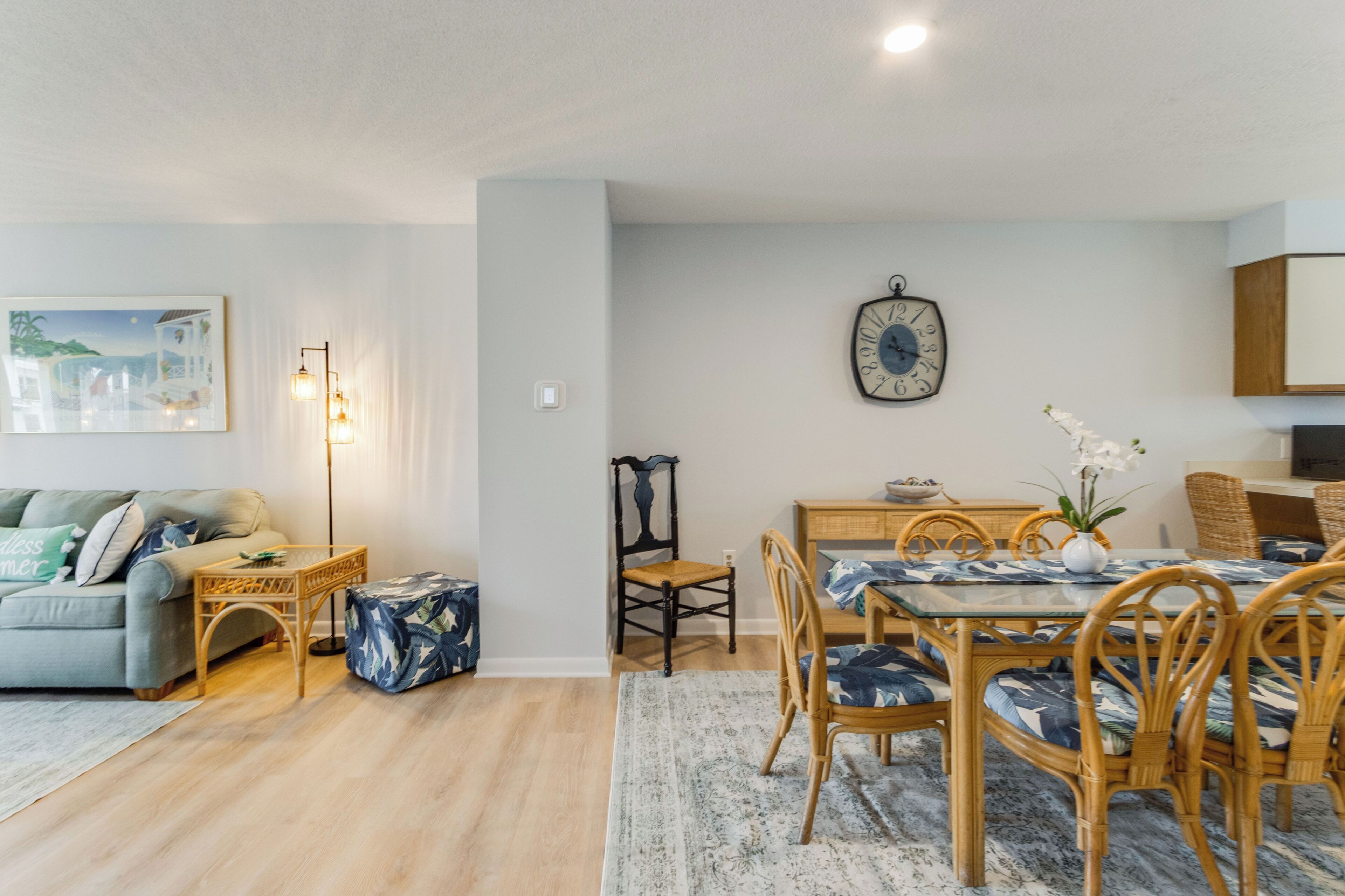A modern living room with a sofa, a dining table with chairs, a side table, a wall clock, and a painting.