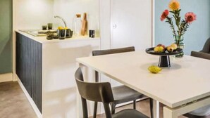 Uma cozinha compacta com bancada branca, fogão embutido e uma pia pequena. Há uma área de refeições com uma mesa branca e quatro cadeiras. Um vaso com flores está sobre a mesa.