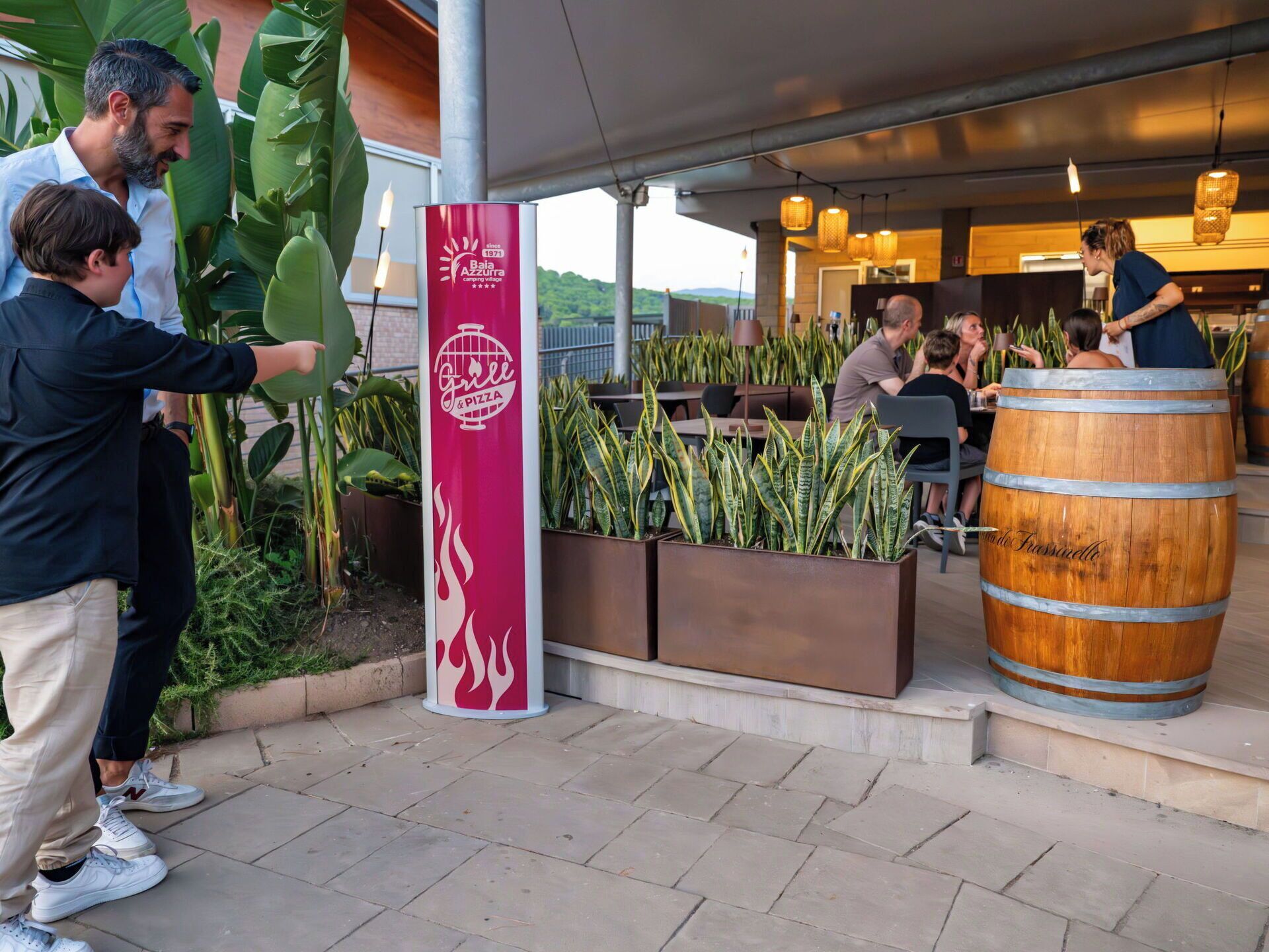 Un espace extérieur aménagé avec des sièges, un panneau rouge indiquant un restaurant, des plantes en pot et un tonneau en bois.