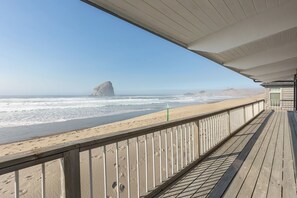 Eine Holzterrasse mit Geländer, die auf einen Strand mit einer im Hintergrund sichtbaren, großen Felsformation blickt.