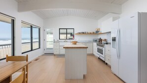 Moderni keittiö, jossa on keskeinen saareke, valkoiset kaapit ja suuret ikkunat, joista avautuu näkymä rannalle.