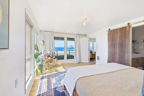 A bedroom with a large bed, a view of the ocean, a sliding door, and a bathroom with a sink and mirror. - Pacific Perch-Lux, 4 bed, oceanfront with huge views. (Pacific City)