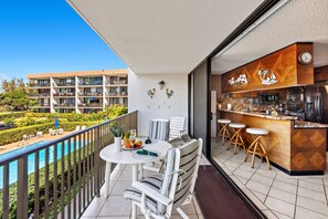 A balcony with a dining table and chairs, a kitchen area with bar stools, and a pool view. - New Listing! Charming Condo Steps to the Beach (Kihei)