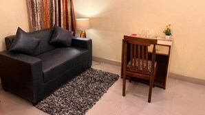 Un salón con un sofá oscuro, cortinas estampadas, una silla de madera, una mesita con una lámpara y un cuadro en la pared.