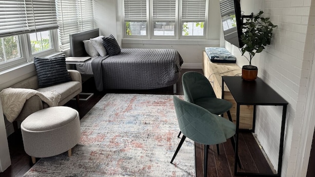 A compact bedroom with a bed, a desk, a chair, a small table, a potted plant, and a window with blinds. - Private Downtown Kitchener Studio | Parking, Kitchenette, Wi-Fi (Kitchener)