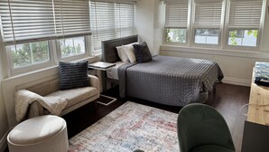 A compact bedroom with a bed, a desk, and a chair. There are windows with blinds, a rug, and a bench. - Private Downtown Kitchener Studio | Parking, Kitchenette, Wi-Fi (Kitchener)