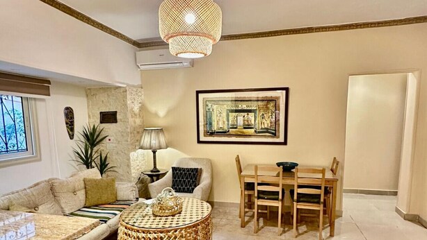 A spacious living room with a round coffee table, a sofa, a dining table with chairs, a large woven lamp, and a framed picture on the wall. - The Helio Suite Modern 1 bdrm (Heliopolis)