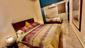 A bedroom with a large bed, a nightstand with a lamp, and a wall-mounted air conditioner. - The Helio Suite Modern 1 bdrm (Heliopolis)