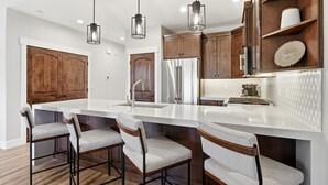 Une cuisine moderne dotée d’un grand îlot central, de tabourets de bar, de meubles en bois, d’appareils électroménagers en acier inoxydable et de luminaires suspendus.