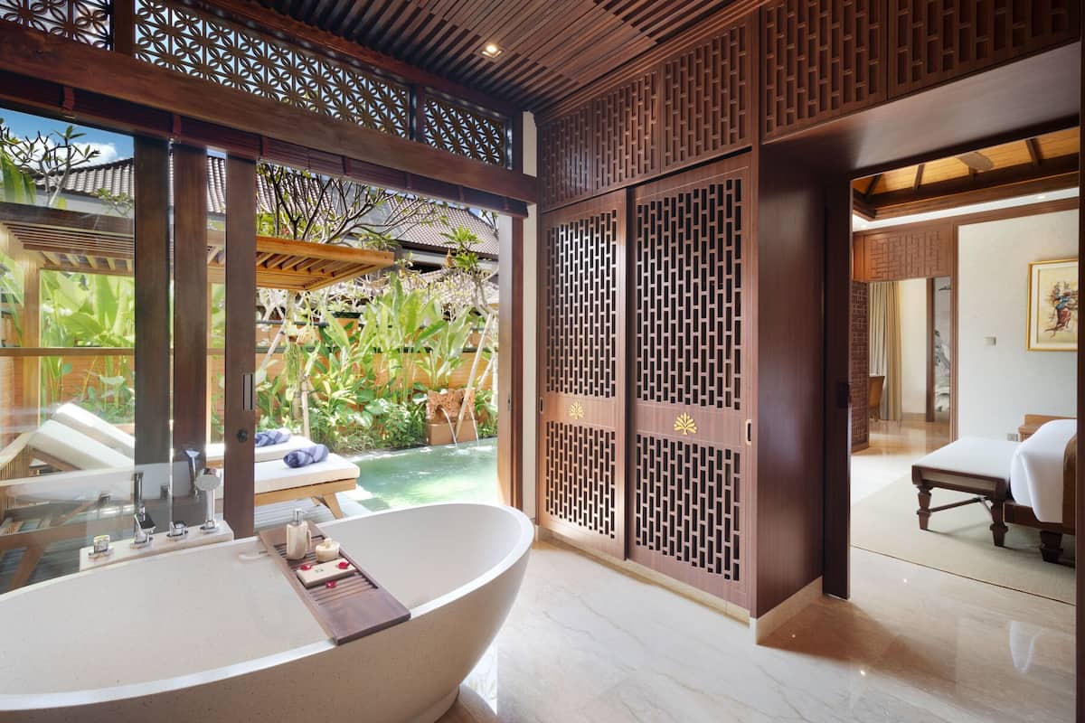 A bathroom with a freestanding bathtub, a sink, and a shower area. There is a wooden partition with a lattice design, and a view of a garden with plants and a pool.