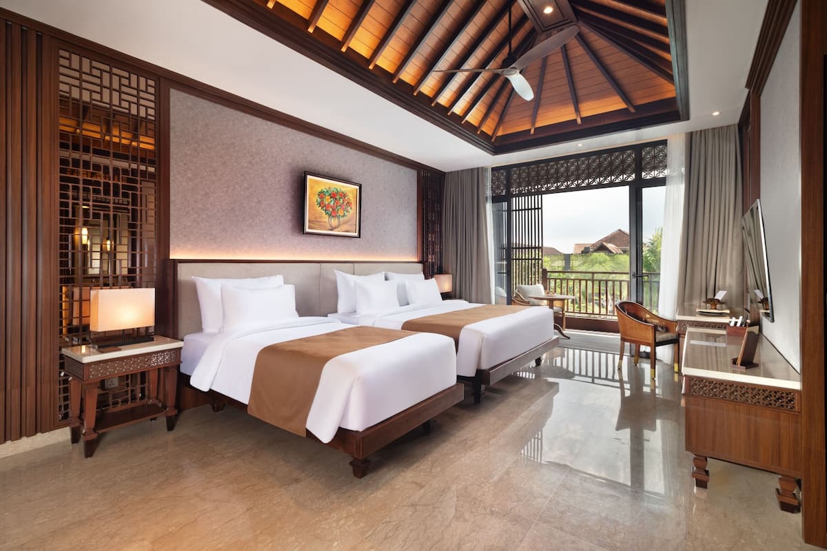 A hotel room with two beds, a desk, a chair, a balcony with a view, and a ceiling fan.
