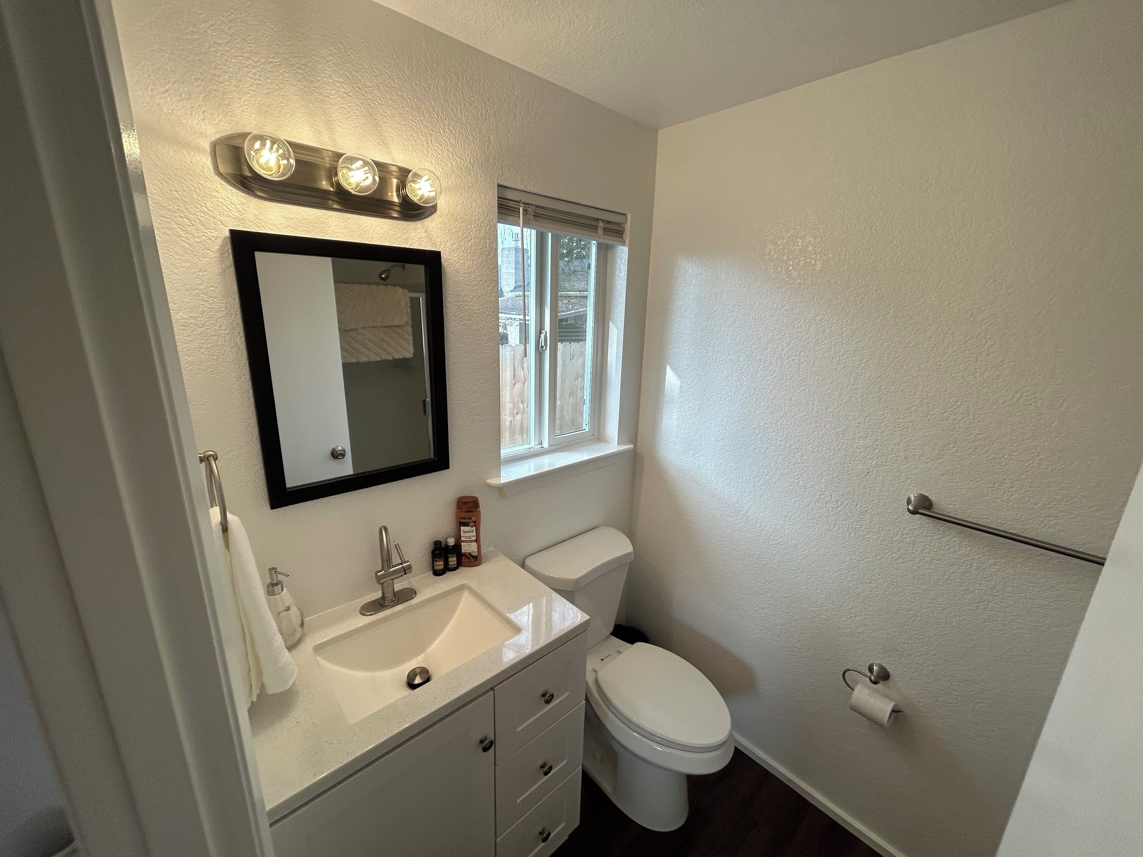 Une salle de bain avec un lavabo blanc, un miroir et des toilettes. Il y a une fenêtre avec des stores, un porte-serviettes et une tringle à rideau de douche.