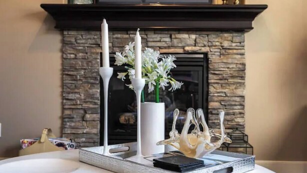 Une table blanche sur laquelle se trouve un plateau portant un vase blanc rempli de fleurs, deux bougies blanches et une sculpture de verre décorative.