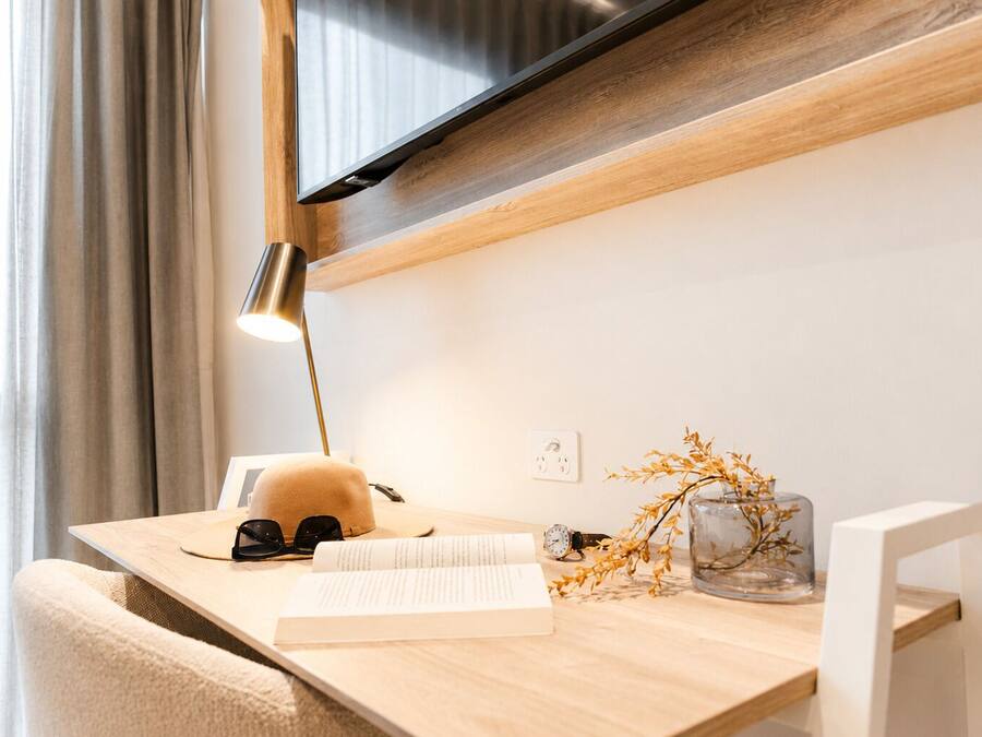 A wooden desk with a lamp, a book, a cap, and a vase with dried flowers.