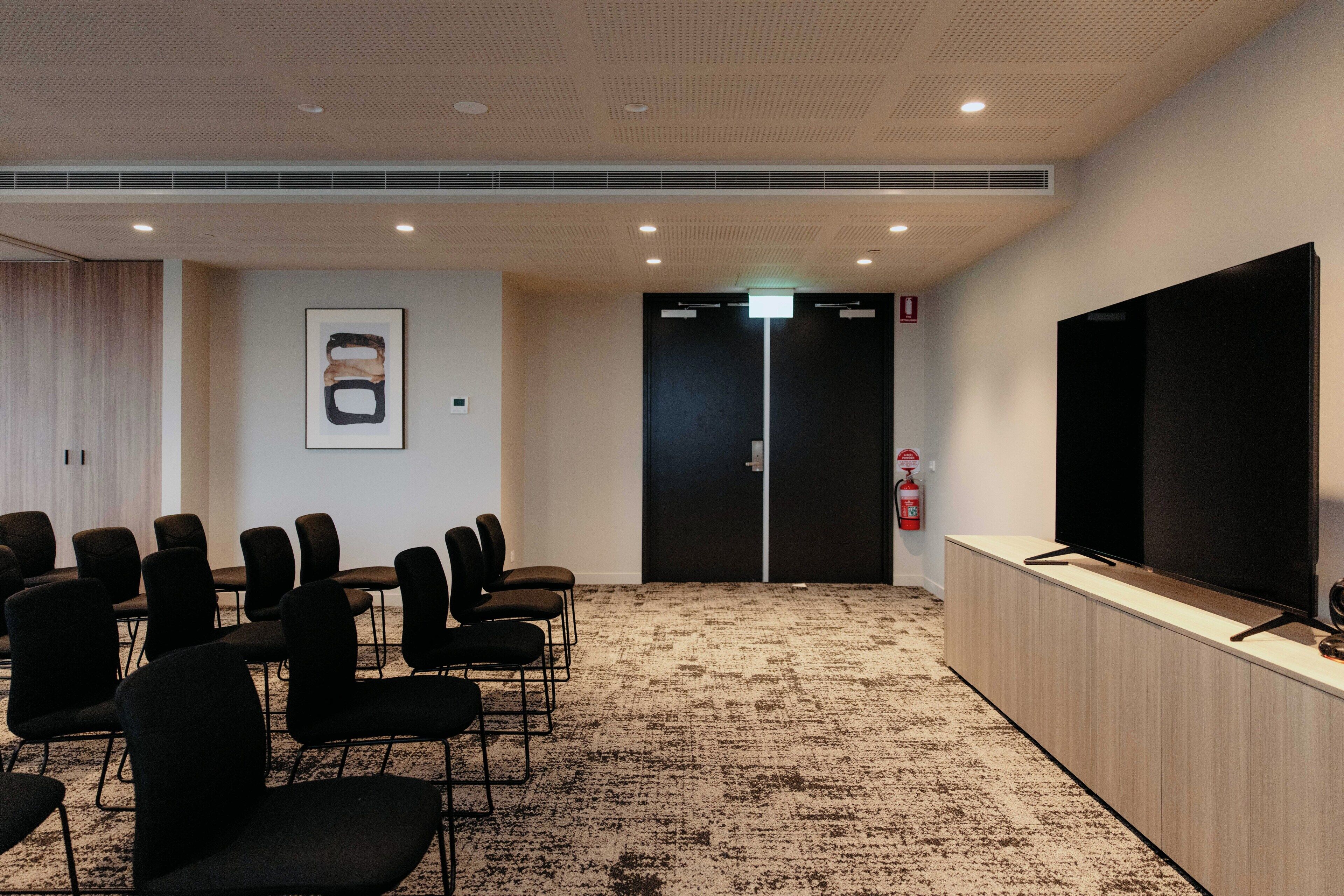 A conference room with a carpeted floor, black chairs, a large flat-screen TV, and a fire extinguisher.