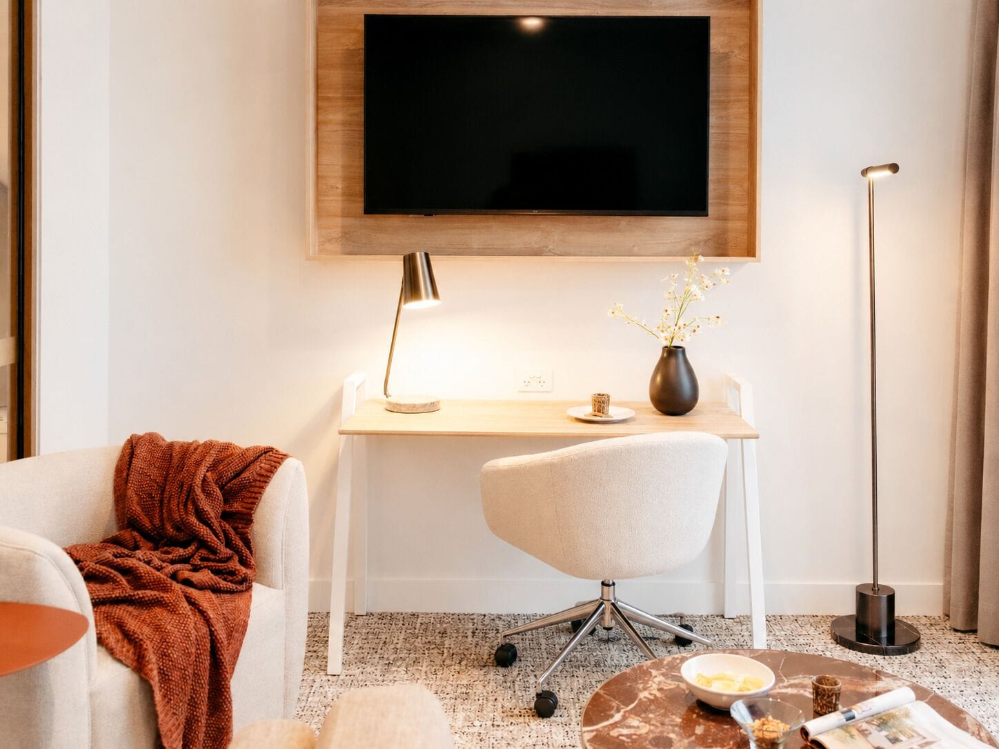Uma sala de estar moderna com uma televisão de tela plana na parede, uma escrivaninha com abajur, uma cadeira, uma mesa de centro com uma tigela e um jornal, e um abajur de chão alto.