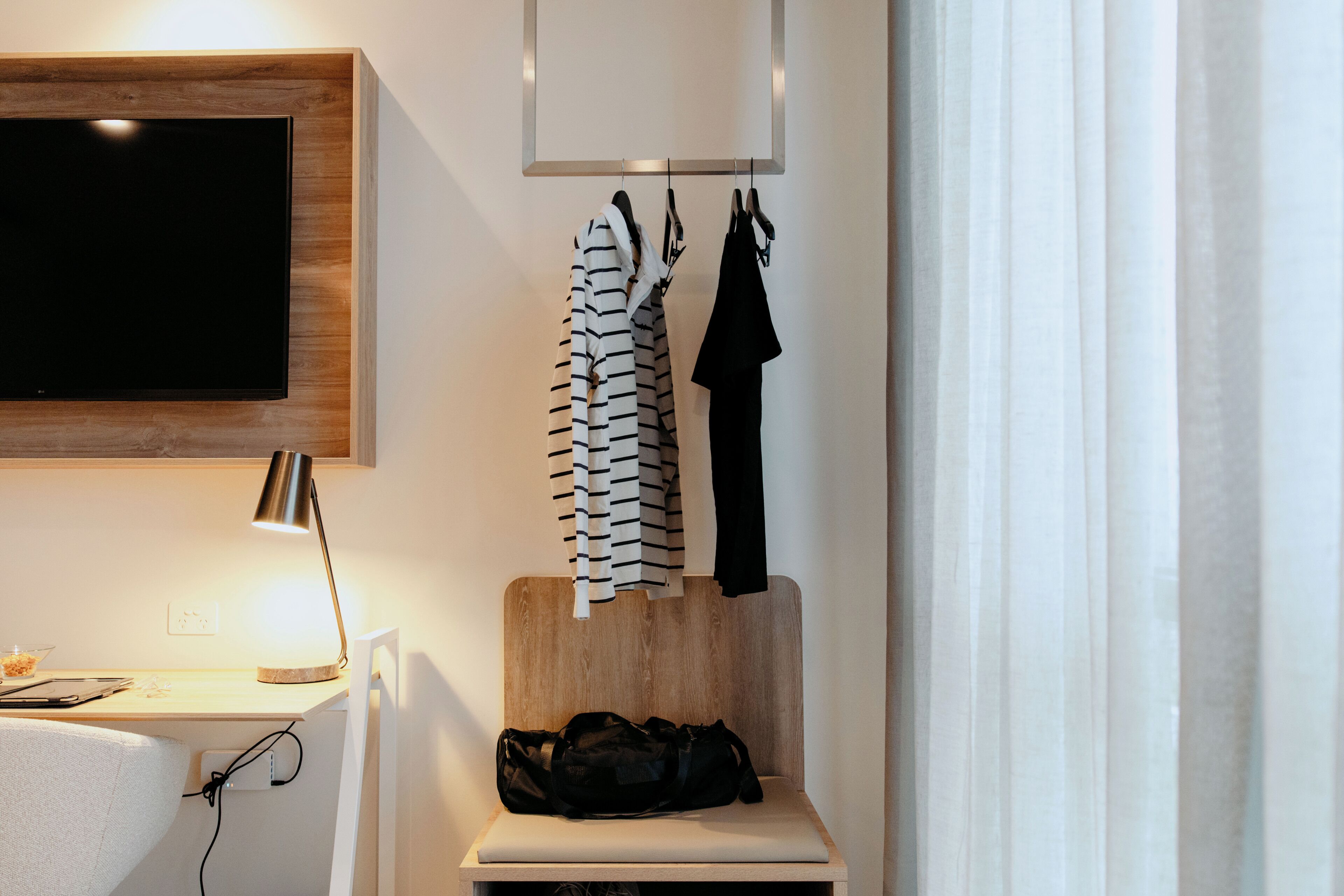 A modern room with a flat-screen TV mounted on the wall, a wooden headboard, a bench with a laptop, and a hanging coat rack.