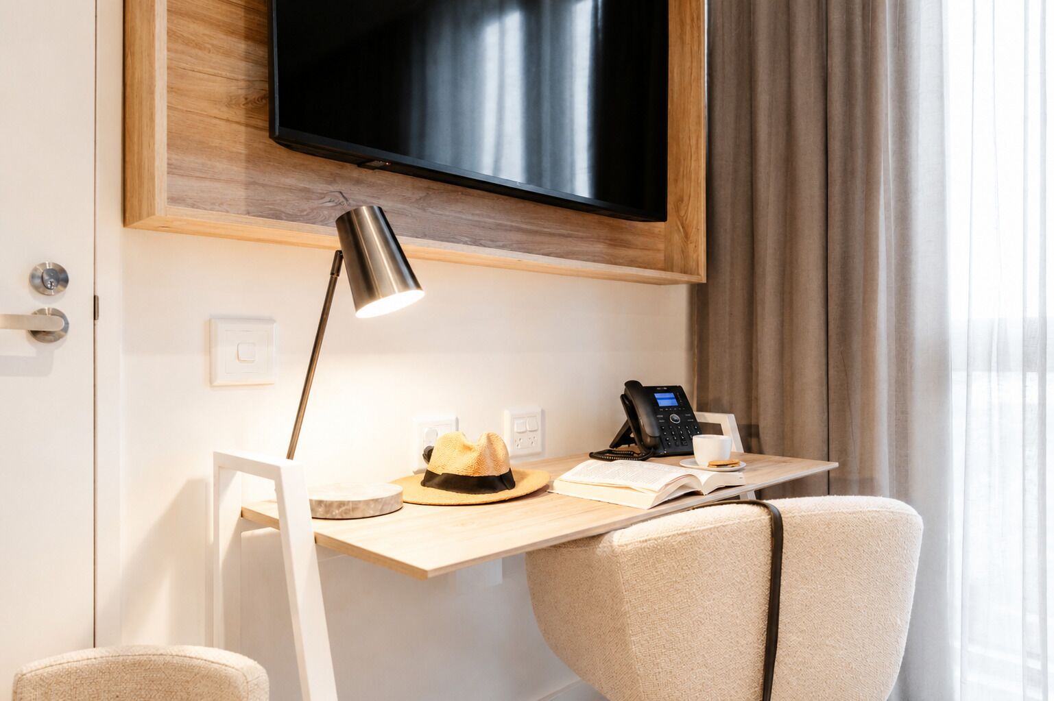 Ein Hotelzimmer mit Schreibtisch, Flachbildfernseher, Schreibtischlampe, Telefon, einem Hut und einem offenen Buch.
