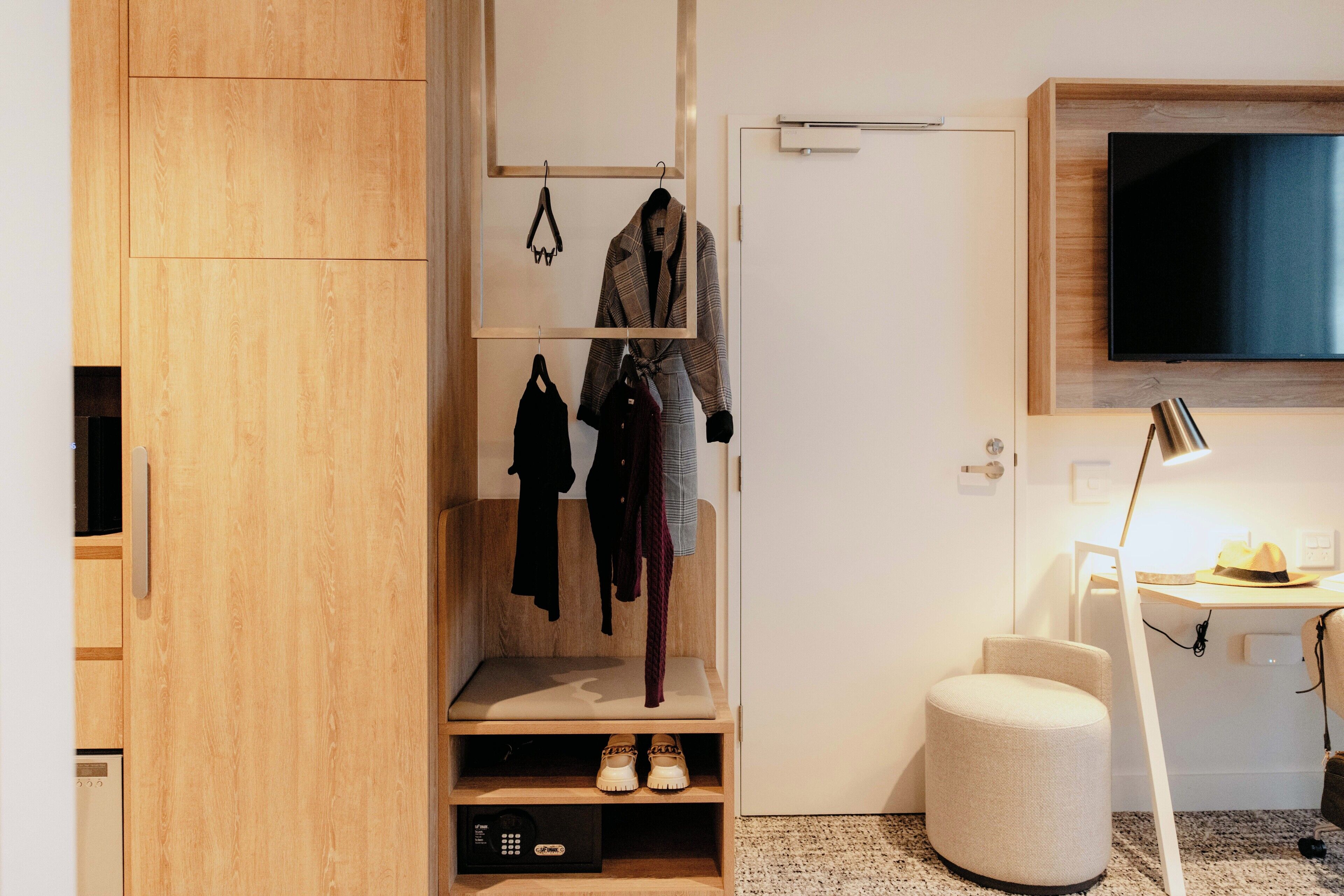 Une chambre moderne avec une armoire en bois, un porte-manteau, une télévision, un bureau avec une lampe et un pouf rond.