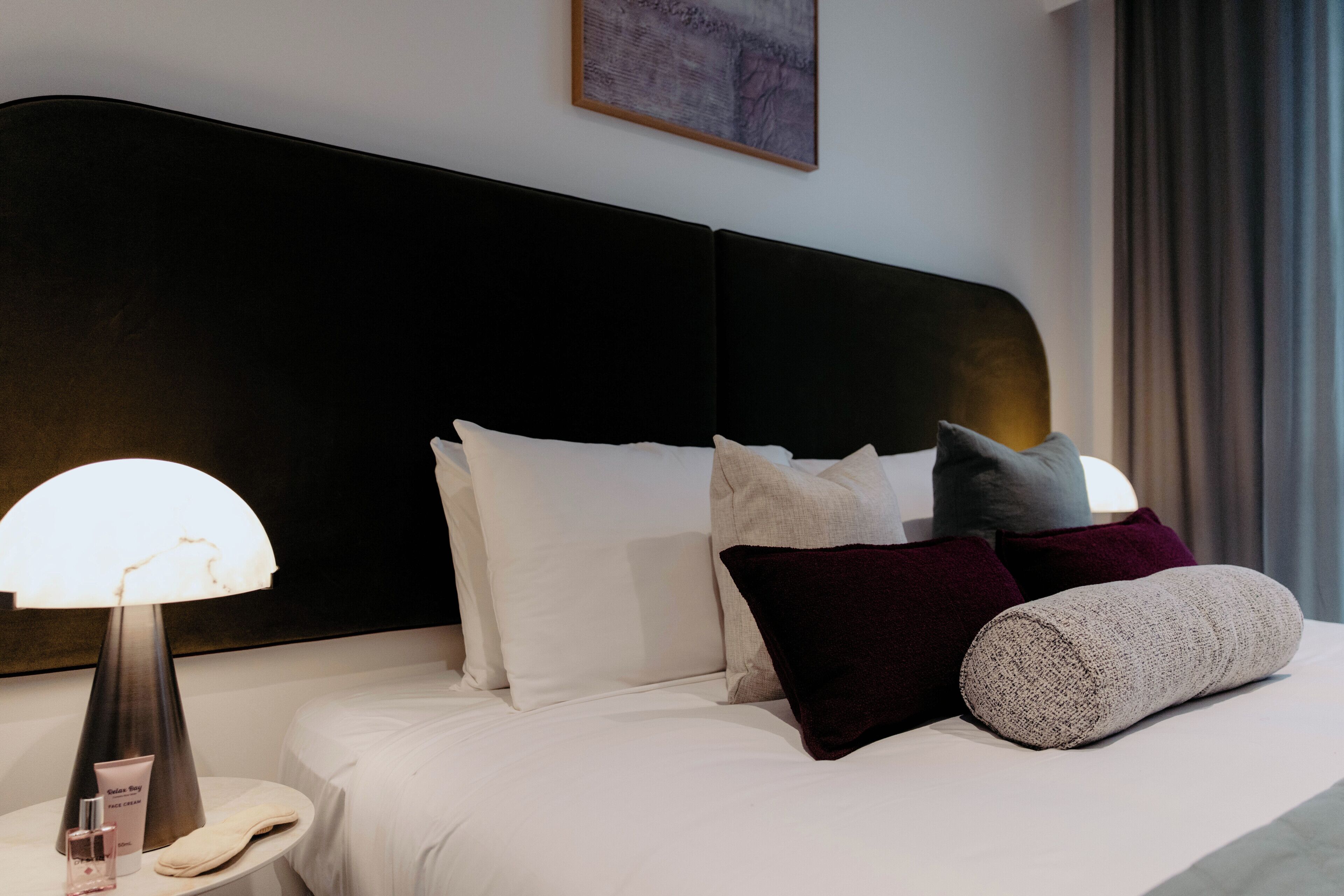 Una cama bien tendida con ropa blanca, una mesita de noche redonda con lámpara y un cabecero oscuro.