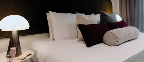 Una cama bien tendida con ropa blanca, una mesita de noche redonda con lámpara y un cabecero oscuro.
